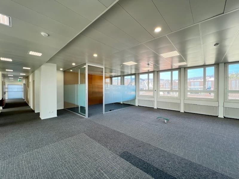 Empty office room with glass walls and windows, tiled floor, white walls, ceiling lights, and pillars.