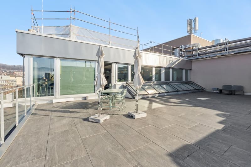 Roof terrace with tiled floor, metal railings, table and chairs, and two closed umbrellas.