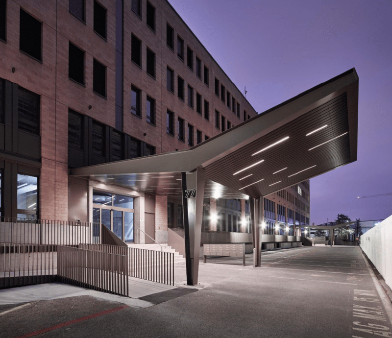 A multi-story commercial or office building with a modern, angular design. The building has a large entrance canopy with lighting, and the exterior is made of concrete and glass. The surrounding area appears to be a parking lot or plaza.