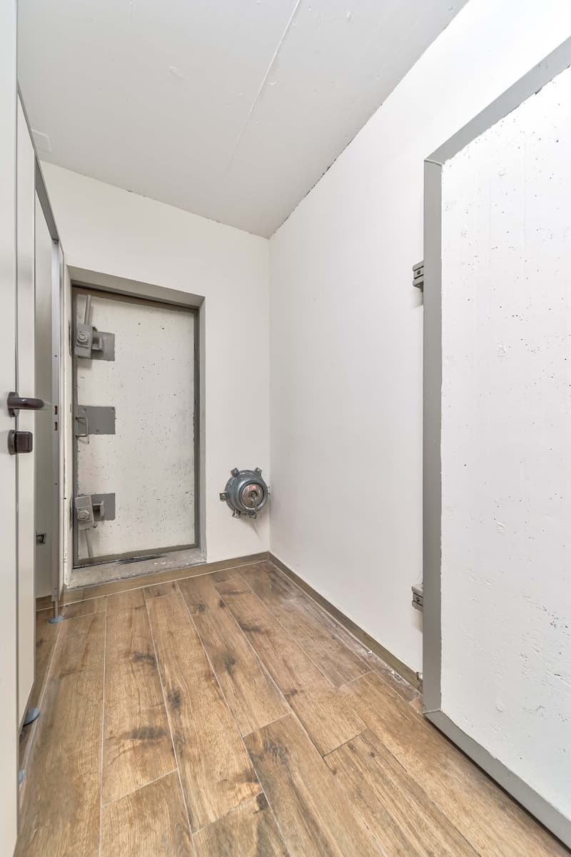 The image shows the entryway of a property. The floor is covered with wood-like tiles, and the walls are painted white. There is a metal door frame and a security camera or other device visible on the wall. The ceiling appears to be flat and painted white