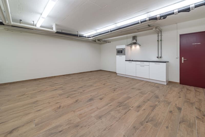 Open-plan studio apartment with hardwood flooring, white walls, exposed ceiling pipes, and a kitchenette with a dishwasher.
