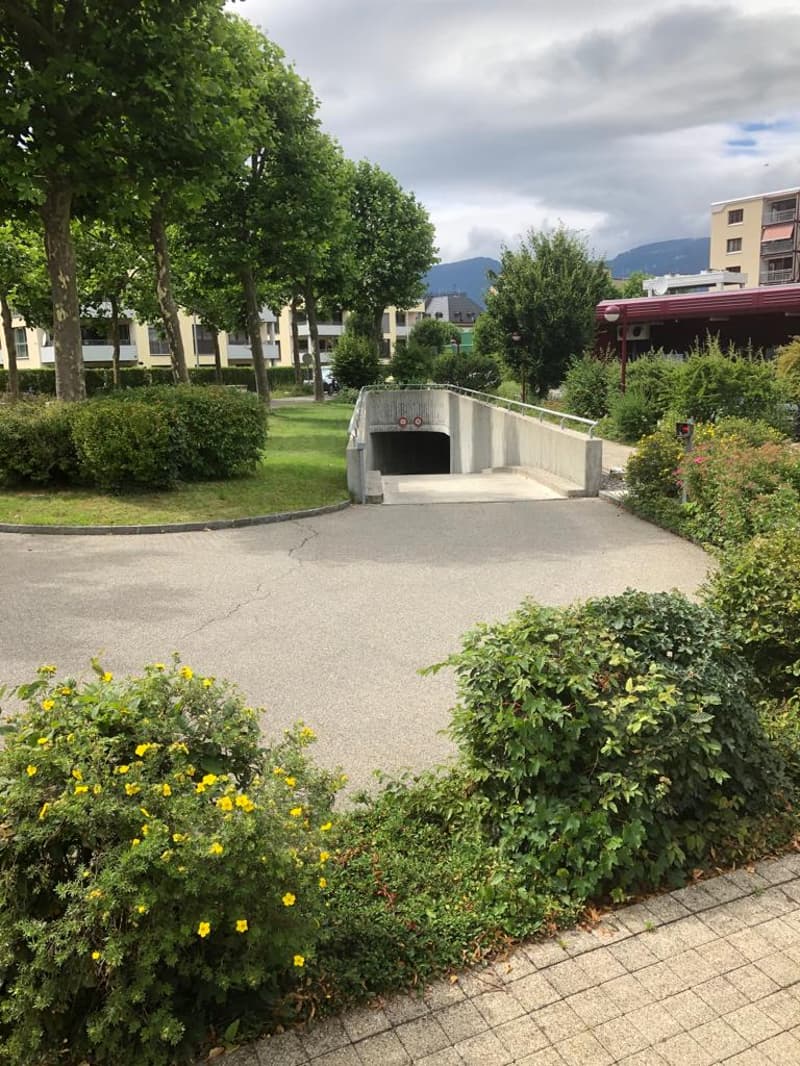 The image shows a well-maintained outdoor area with lush greenery, including trees, shrubs, and flowers. There is a paved walkway leading to a covered parking area or tunnel. The surrounding buildings and mountains can be seen in the background, suggestin
