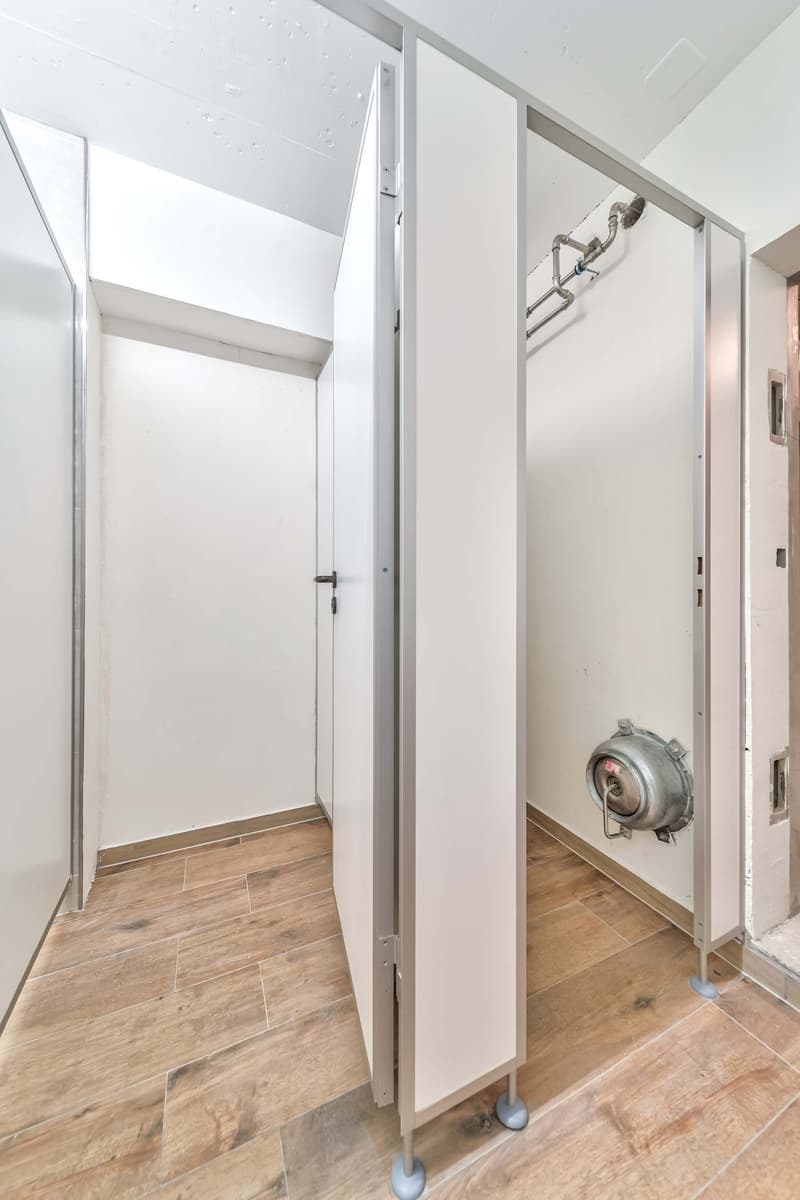 The image shows a small laundry room or utility closet with a washing machine visible. The room has white walls and a tiled floor with a wood-like pattern. There is a metal door frame and a metal appliance, likely the washing machine, can be seen in the c
