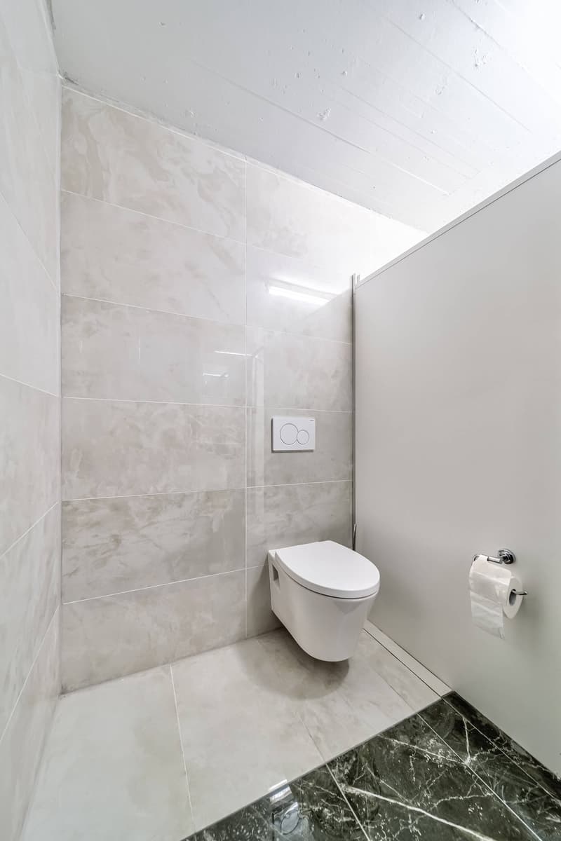 The image shows a modern bathroom with white tile walls and a black marble floor. The bathroom features a wall-mounted toilet and a toilet paper holder. The room appears to be well-lit with a clean and minimalist design.