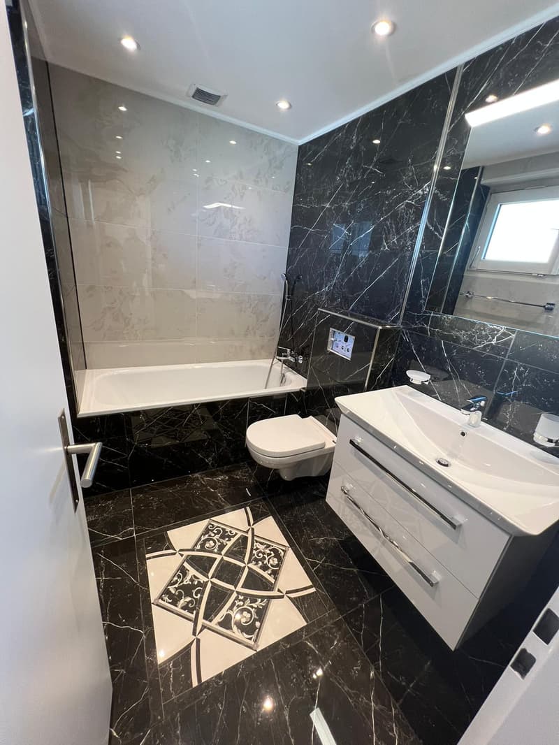 modern bathroom with black and white marble tiles, white bathtub, modern white toilet, white sink with two drawers, mirror with built-in light