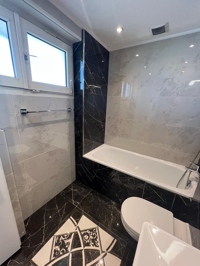 Marble tiled floor, white and black tiled walls, bathtub with showerhead, white toilet, white sink, towel rack