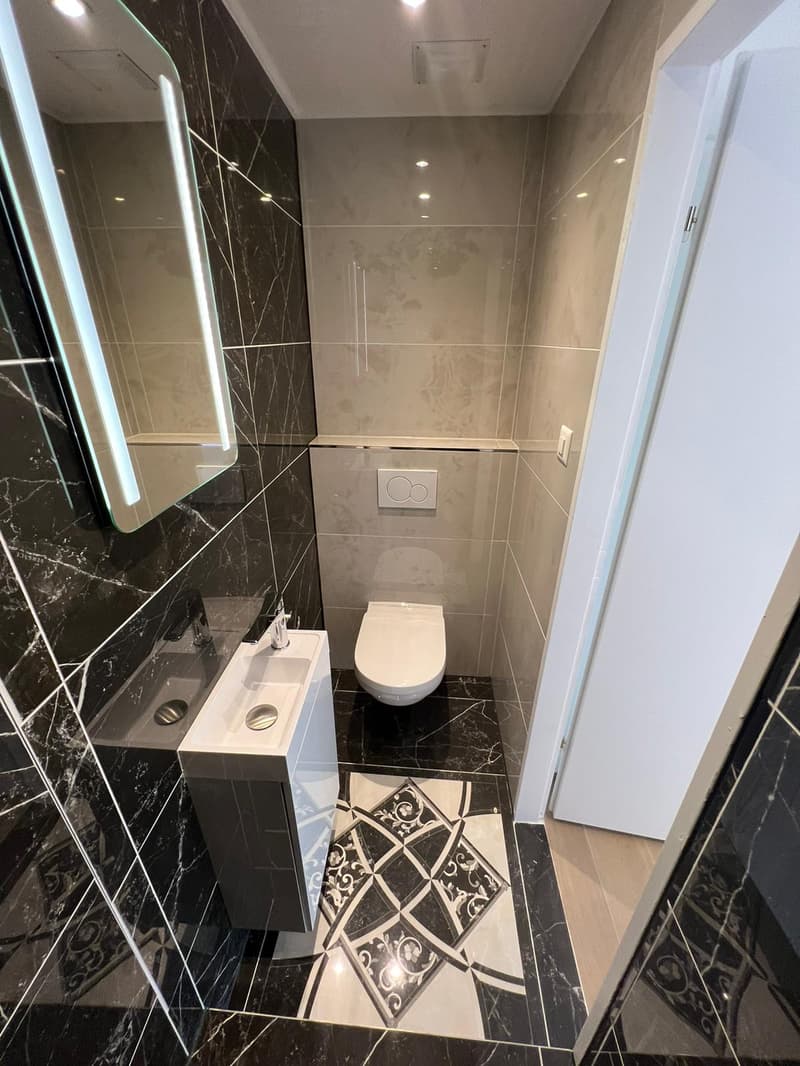 Modern bathroom with black and white marbled tiles, a small white pedestal sink, a toilet, a small mirror, a white door