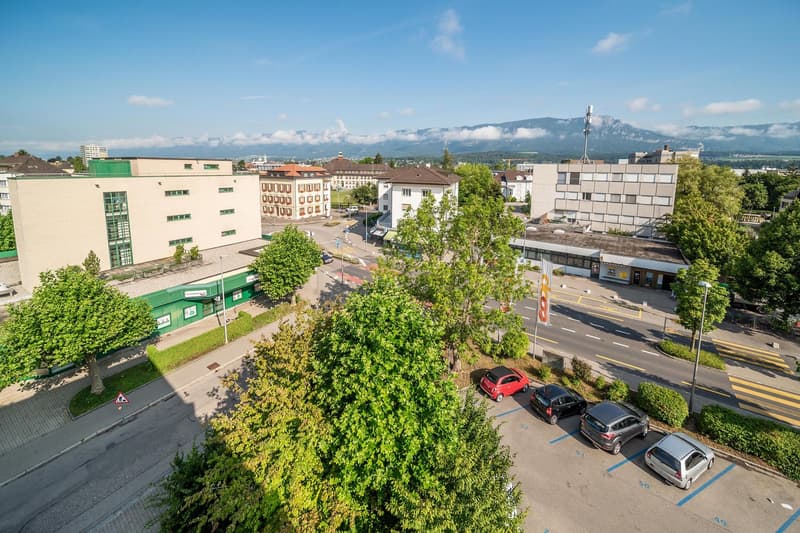 Several buildings with parking lots, cars parked, trees, road with traffic signs, mountains in the distance
