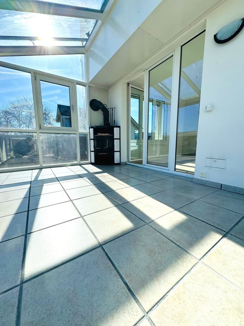 A spacious balcony with a tiled floor, large glass doors, and a wood stove. The balcony has a view of the outside with trees and a house.