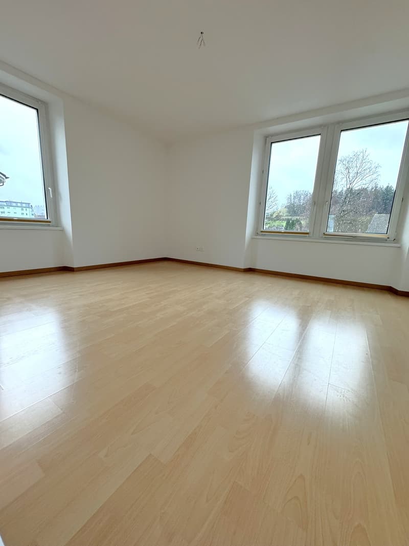 Large, empty room with hardwood flooring, white walls, and windows that provide a view of the outdoors.