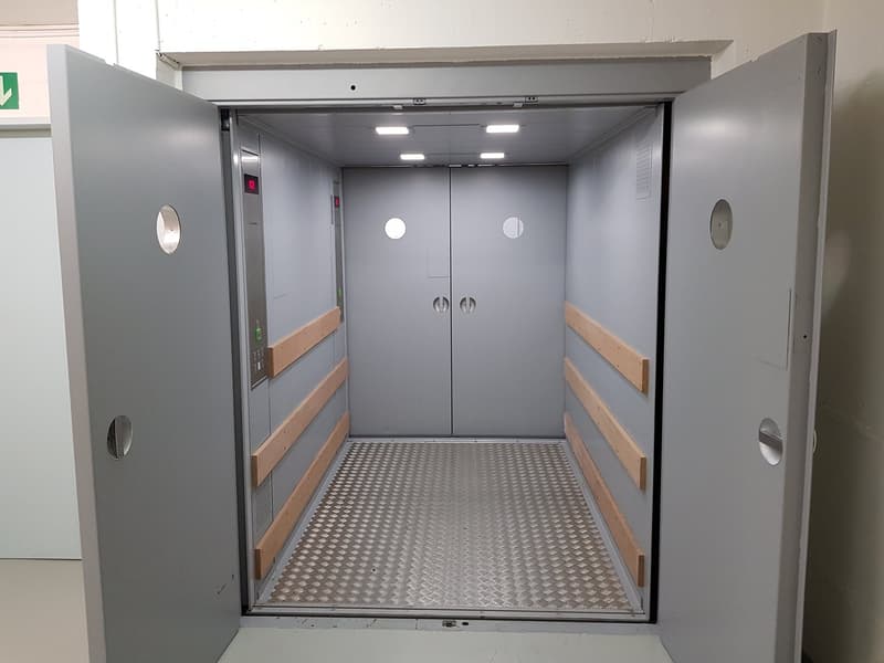 The image shows the interior of an elevator. The elevator has a gray metal frame and double doors. The floor is made of a metal grate material. The interior is well-lit with LED lights.