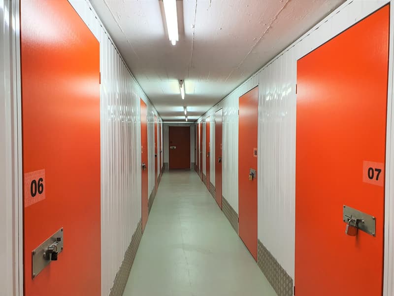 The image shows a long, narrow hallway with numbered storage units on either side. The walls are painted in a bright orange color, and the floor is a plain concrete. The hallway appears to be part of a storage facility or warehouse, with the numbered unit