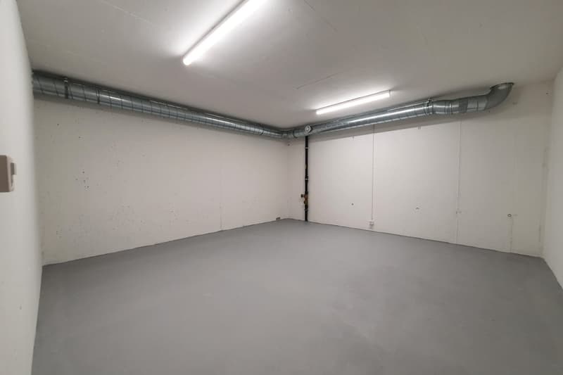 This image shows a large, empty garage space with a concrete floor and white walls. The ceiling is equipped with ventilation ducts and lighting fixtures, indicating this is a functional garage or storage area.