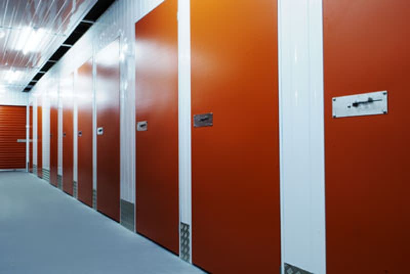 The image shows a row of orange storage units or lockers in a commercial storage facility. The units have metal doors and appear to be in a well-lit, industrial-style space with a concrete floor.