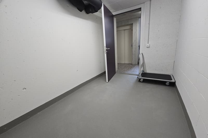 an empty room with concrete floor, white walls, a door to another room, and a dolly in the corner