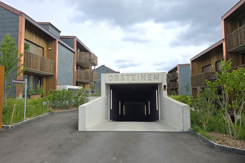 A modern underground parking garage with concrete entrance, 'Obsteinen' sign above, concrete walls, and metal railings. Adjacent multi-story buildings with balconies and greenery.
