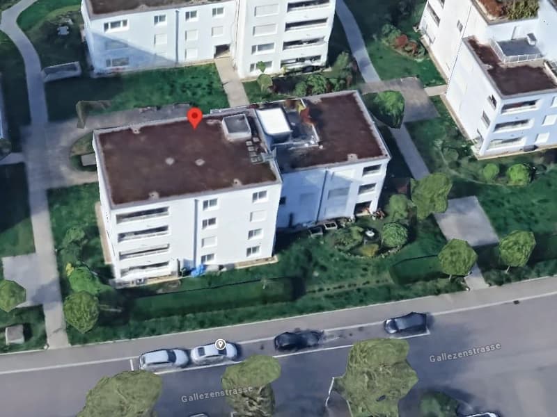 Aerial view of a white building with multiple floors, surrounded by trees and grass, with a red marker indicating a specific location.