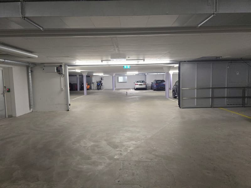 Empty parking lot with concrete floor, white walls, metal beams, and fluorescent lights. Several cars parked in designated spots, and a motorcycle parked in a corner.