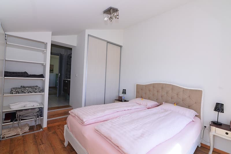 Bedroom with two beds, white walls, wooden floor, white wardrobe, shelves, bedside tables, lamps, and a nightstand.