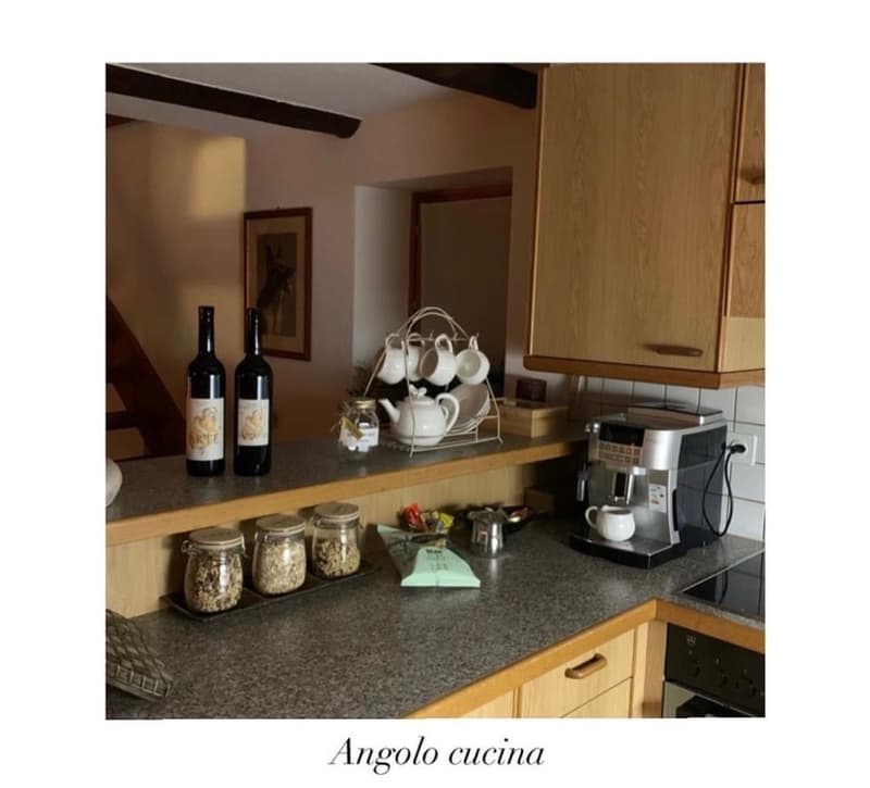 The image shows a cozy kitchen with wooden cabinets, a granite countertop, and various kitchen appliances and accessories. There is a coffee maker, jars with dry goods, and a display of teacups and a teapot. Two bottles of wine are also visible on the cou