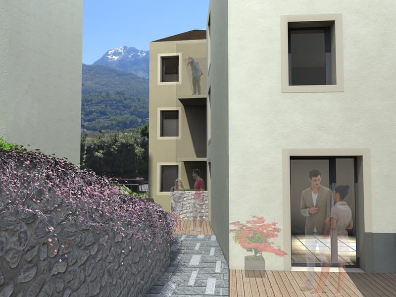 A modern apartment building with large windows and balconies overlooking a scenic mountain landscape. The building has a stone wall and wooden deck elements. There are people visible on the balconies.