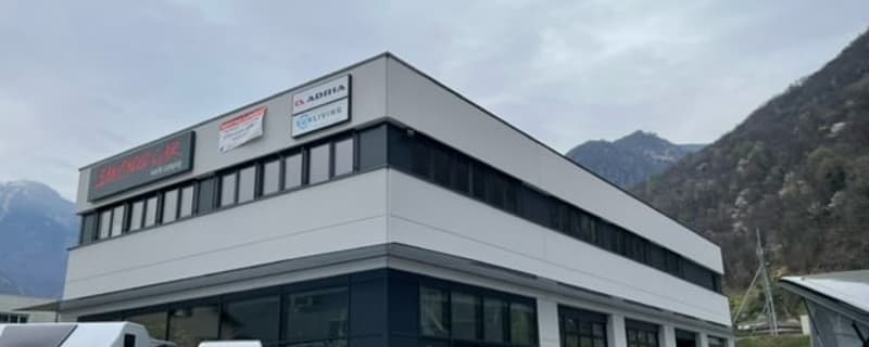A multi-story commercial building with a large parking area in front. The building has a modern, industrial-style design with a mix of glass and metal panels. The building appears to be used for some kind of commercial or industrial purpose, based on the