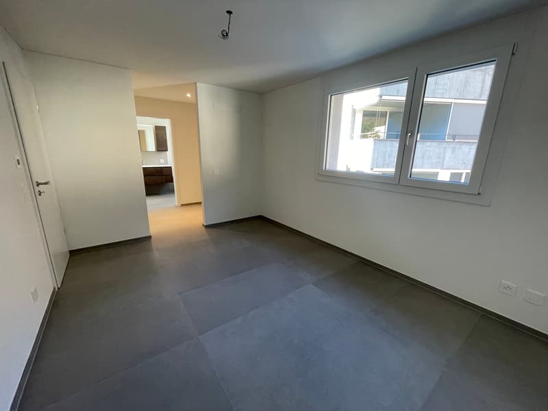 Empty room with white walls, tiled floor, glass windows, visible doorway to the hallway and a bathroom sink and mirror in the hallway.