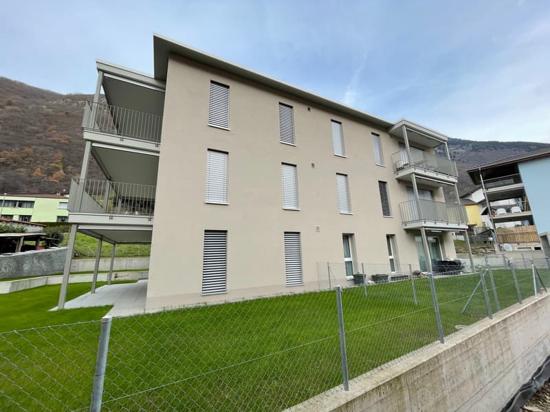 3-story apartment building with beige exterior, balconies, and large windows. The building is surrounded by a green lawn and fenced area.