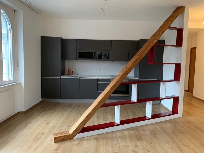 Modern kitchen with black cabinets, white walls, wooden beams, and a red and white shelving unit next to the kitchen.