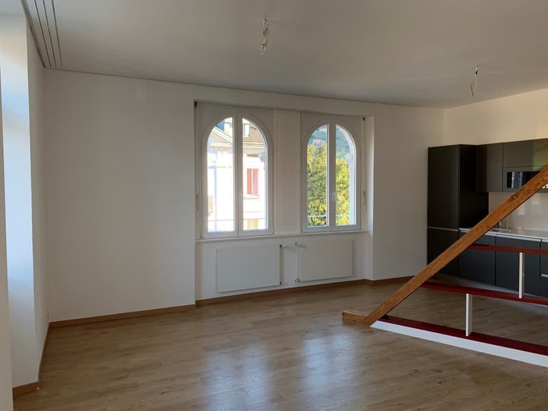 Spacious living room with large arched windows, hardwood floors, and a modern kitchen area with black cabinets.