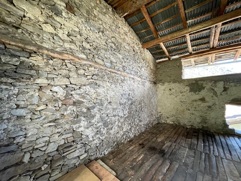 The image shows the interior of an old, rustic building. The walls are made of rough, uneven stone, creating a rugged, natural appearance. The ceiling is constructed with wooden beams and corrugated metal roofing. The floor is made of wooden planks. The o