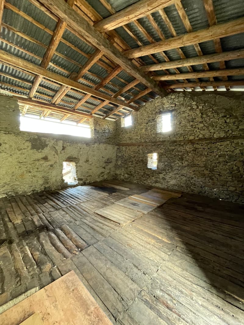 The image shows the interior of an old, rustic building. The walls are made of stone, and the ceiling is constructed with wooden beams and corrugated metal roofing. The floor is made of weathered wooden planks. The space appears to be a large, open room w