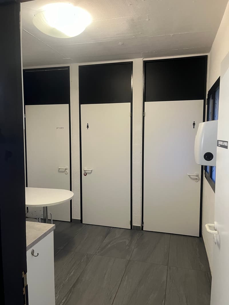 Empty bathroom with white tiles, white walls, three stall doors, table, dispenser