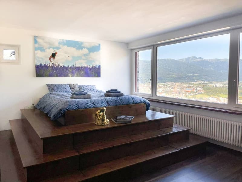 Wooden platform with bed, books and statue, large windows, painting on wall, wooden floor