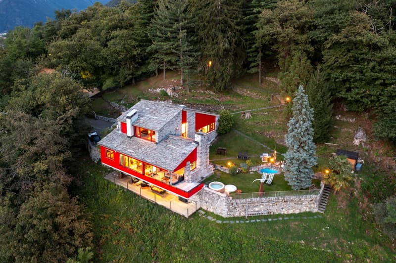 Rustic house with stone walls, red facade, gray roof, swimming pool, large windows, patio with outdoor furniture, surrounded by lush greenery and trees