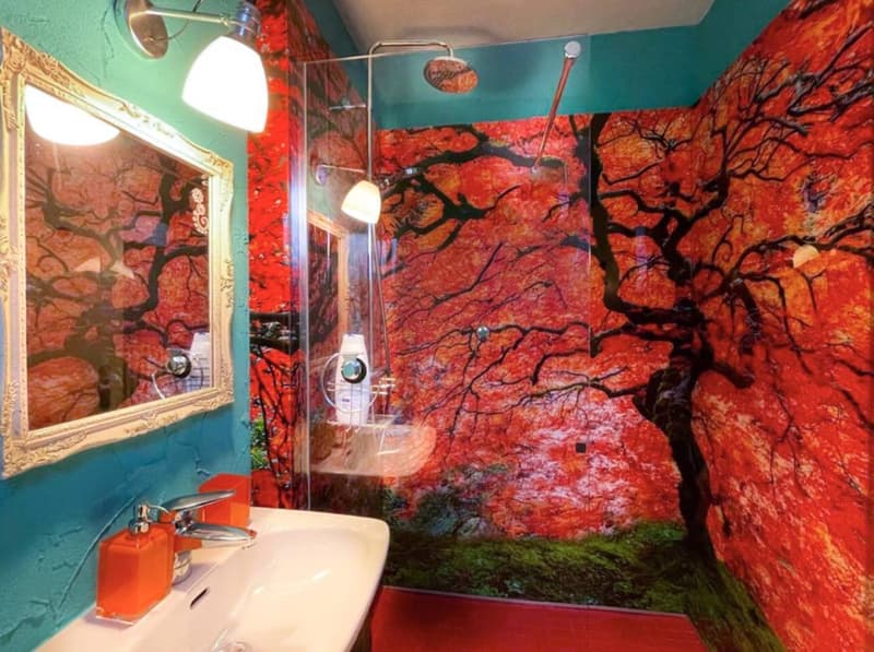 A bathroom featuring a white sink, a mirror, a glass shower cubicle, a showerhead, and walls with a tree mural in red and green colors.
