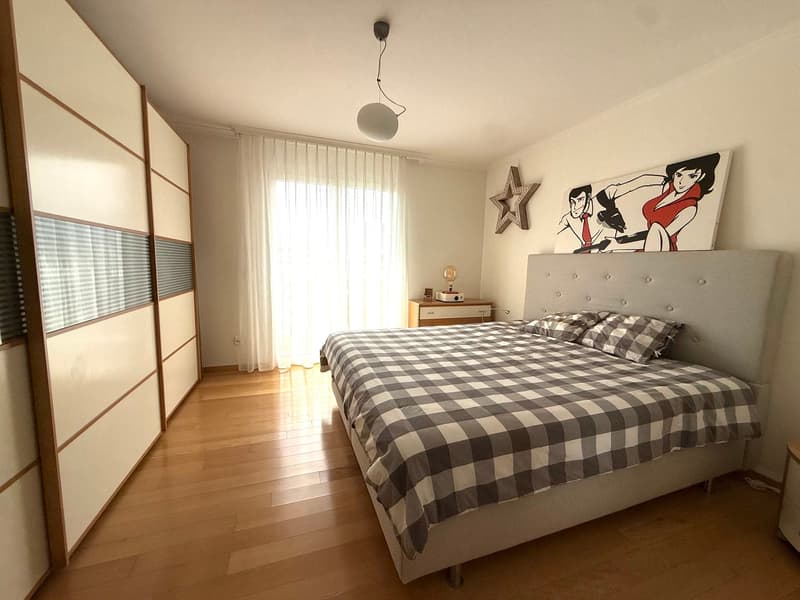 Bedroom with wooden floor, white walls, bed with checkered bedspread, nightstand with lamp, and a painting on the wall.