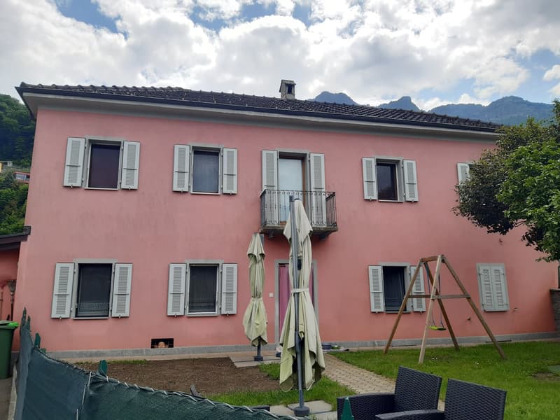 A two-story pink building with white shutters and balconies, surrounded by trees and mountains in the background. There is a ladder and some outdoor furniture in the foreground.