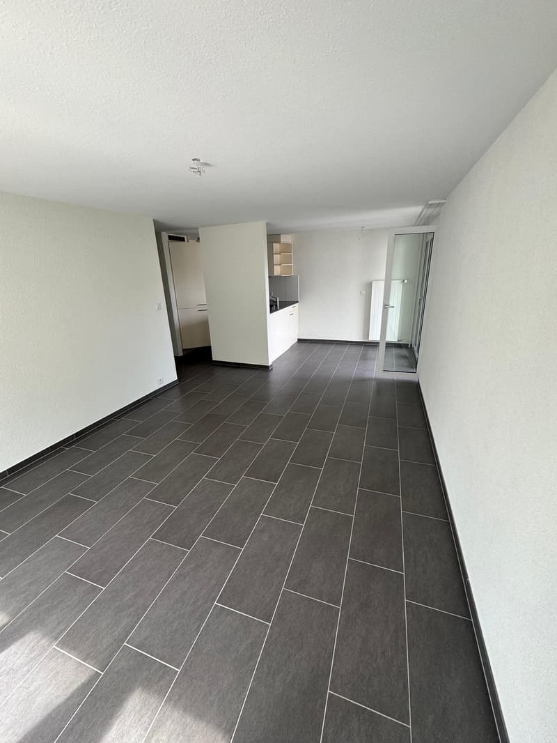 an empty room with tiled flooring, white walls, an open door leading to a kitchen, and a small balcony visible through a glass door