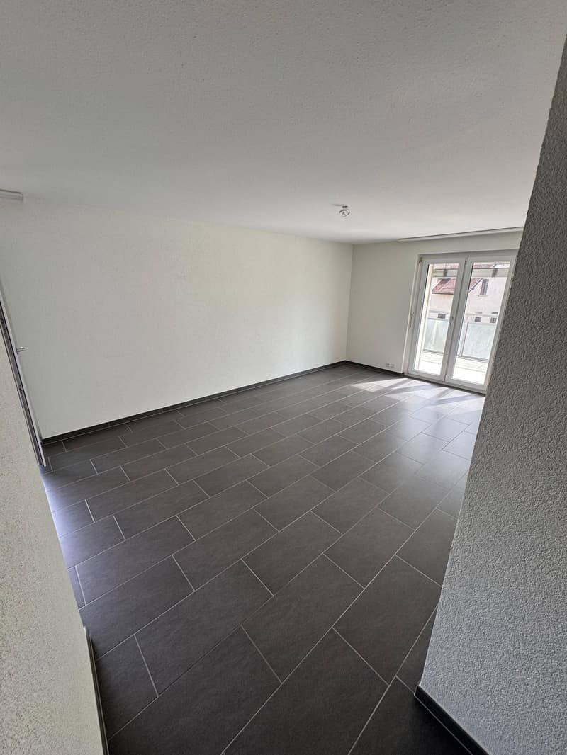Large empty room, modern flooring, white walls, sliding glass doors leading to balcony, power outlet on wall