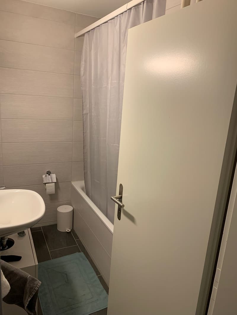 Bathroom with white tiles, white sink, white bathtub, white shower curtain, white toilet, white trash can, blue rug, towel on a towel rack
