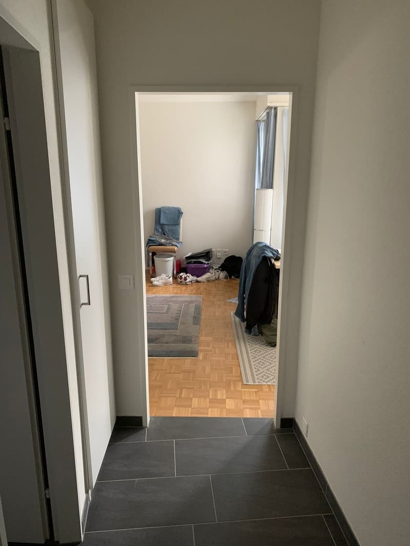 Empty room with wooden floor, white walls, open door, rug, chair, and various items on the floor