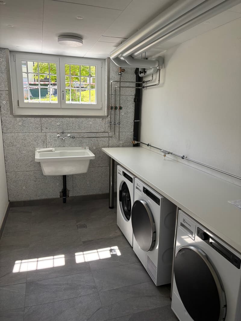 Laundry room with washing machines, sink, and pipes. Tiled floor and white walls.