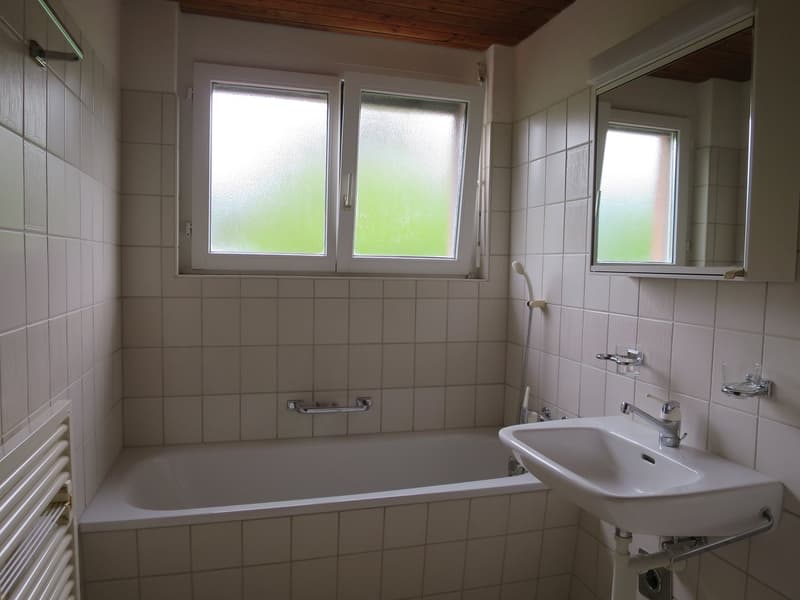 A white bathroom with a bathtub, sink, shower attachment, mirror, and white tiled walls.