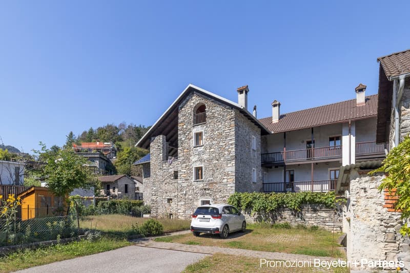 Stone house with multiple balconies, chimneys, white car parked in front, pathway leading to the entrance
