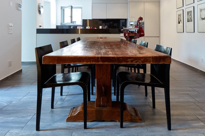 Dining room with a large wooden table and black chairs, white walls, tiled floor, and a kitchen in the background.