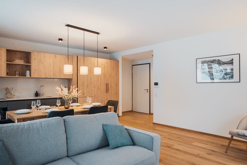 The image shows an open-plan living room and kitchen area. The kitchen features wooden cabinets and shelves, a black countertop, and a dishwasher. The living room has a gray sofa and a large framed artwork on the wall. The room has a modern, minimalist de