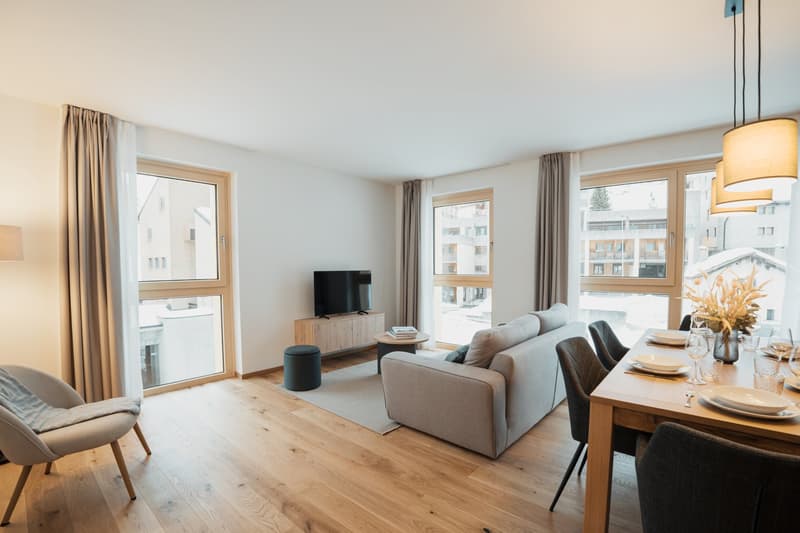 Spacious open-plan living room and dining area with large windows, hardwood floors, gray sofa, wooden TV stand, and dining table with chairs. The room has a cozy and modern feel.
