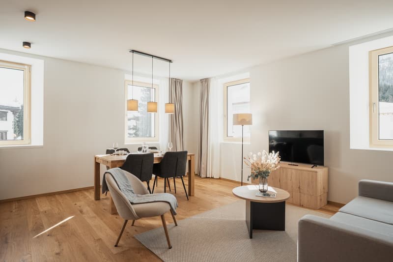 Spacious living room with hardwood floors, large windows, dining table, chairs, sofa, TV stand, and pendant lighting fixtures