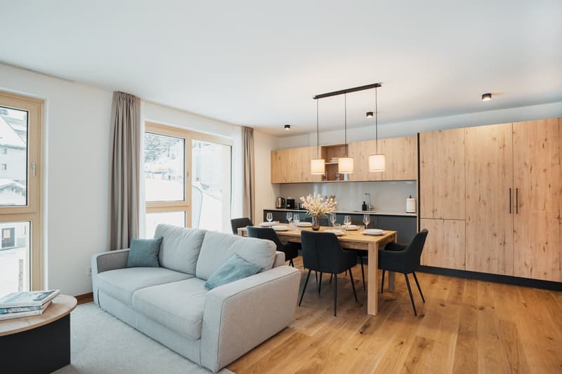 The image shows an open-plan living room and kitchen area. The living room has a comfortable gray sofa with throw pillows. The kitchen features wooden cabinets, a black countertop, and a dining table with black chairs. The room has large windows that prov